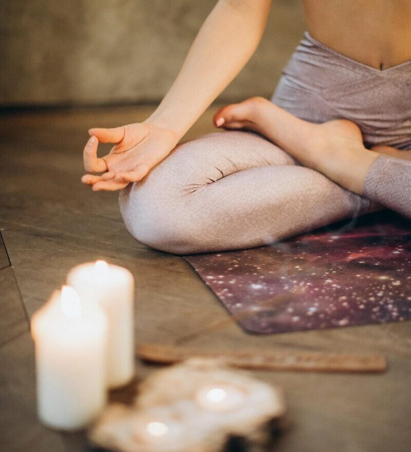 A woman practicing yoga indoors in lotus pose with candles creating a calming atmosphere.